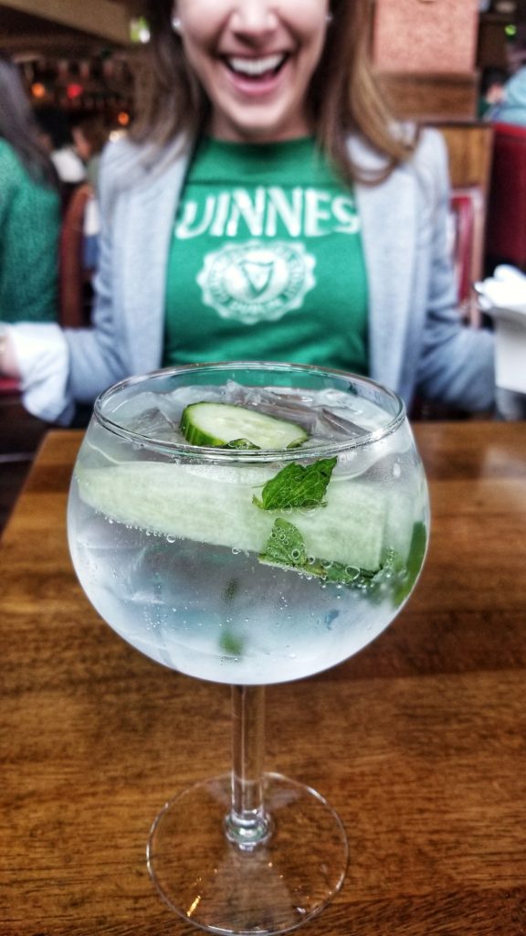 Pride of the Peninsula cocktail in goblet with ice and long strips of cucumber with fresh mint on table. Green Guinness Tshirt on smiling lady in background.