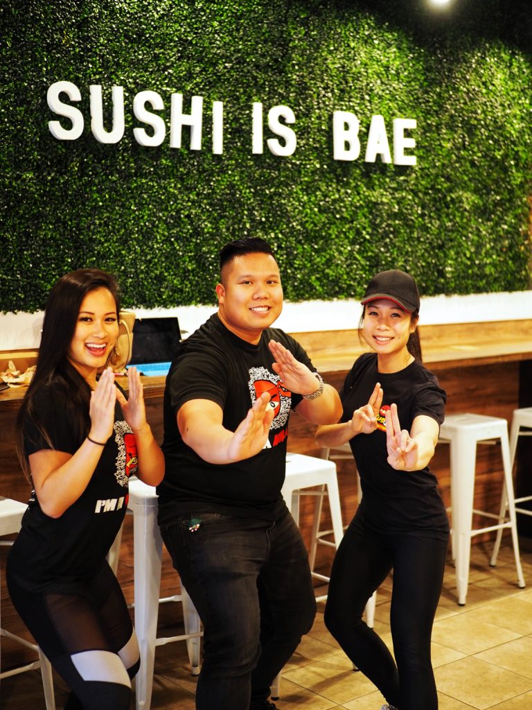 Three Sus Hi employees (Tiffany Nguyen, Milton Tu and Tiffany Chan) strike their best ninja poses. The trio is standing in front of a green wall with Sushi Is Bae during the media preview of the new Sus Hi Colonial Drive location in Mills 50 of Orlando, FL. Taken by Lisa Wilk of TasteCookSip